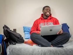 Photo of student using laptop while sitting on bed in residence hall.